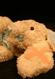 Photo from the programme of the play Playing God by Paula Garfield and Rebecca Atkinson: a toy cuddly rabbit is down with an injury on one ear as shown by a cross of bandages.